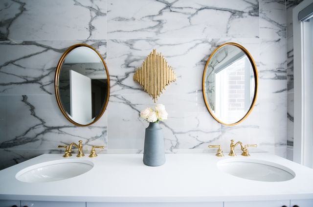 Los mejores accesorios de baño para su hogar compartido, double vanity mirrors outlined in gold on the marble wall with double sinks under the mirrors.